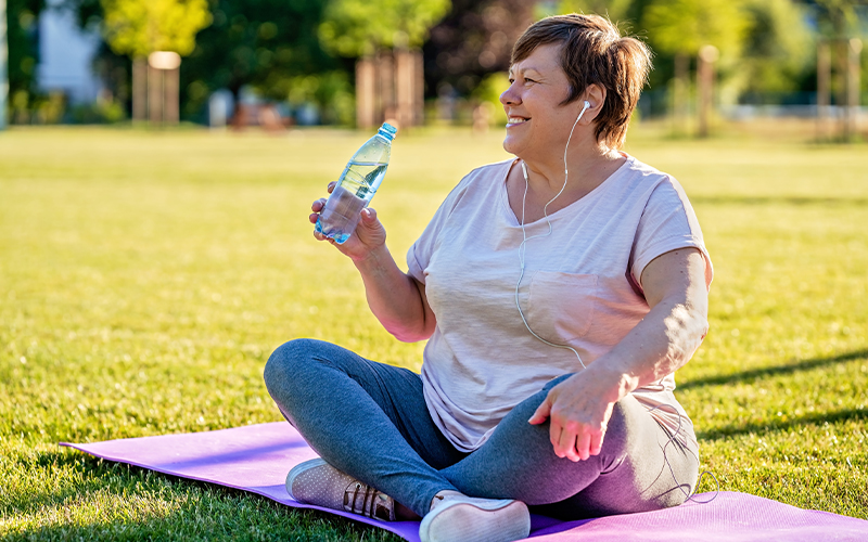 A woman sitting on a yoga mat in the grass drinking water